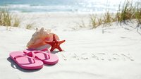 Pink flip flops, conch shell, and starfish on white sand beach with vacation text written in sand, sunny summer holiday concept.