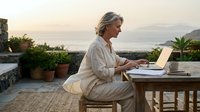 Mature woman with gray hair working on laptop at outdoor terrace table with mountain and sea views, remote work and freelance lifestyle concept for business and productivity themes.