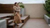 Young Caucasian mother in beige dress holding toddler daughter on modern outdoor wooden deck terrace with contemporary landscaping and planters.