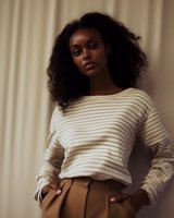 Young African American woman with natural curly hair wearing striped cream sweater and brown pants poses confidently against neutral curtained background in warm lighting.