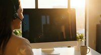 Young African American woman with headset working on computer at bright office desk during golden hour sunrise creating warm atmospheric lighting for remote work concepts.
