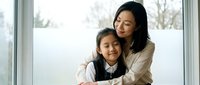 Asian mother and daughter sharing a warm embrace near a bright window, expressing love, care, and family bond in a cozy home setting.