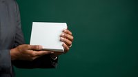 Person with dark skin tone holding blank white card or paper against green background, suitable for mockups, presentations, and advertising campaigns.