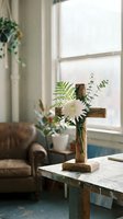 Wooden cross decorated with white chrysanthemum and green ferns on rustic table near window in cozy living room with natural light and houseplants.