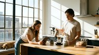 Young couple cooking together in modern kitchen with natural light streaming through large windows creating warm domestic atmosphere.