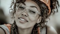 Young mixed-race female construction worker wearing safety glasses and hard hat smiling confidently at camera in industrial workplace setting.