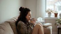 Young woman with dark hair and olive complexion sitting on beige sofa, holding warm beverage mug, relaxing at home near window with natural light and potted plants.
