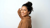 Young Hispanic woman with curly hair in messy bun wearing brown shirt smiling at camera with hand near face against white background for lifestyle content.