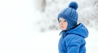 Young child wearing blue winter hat with pom-pom and matching jacket, looking away in snowy outdoor setting. Suitable for seasonal campaigns and family content.