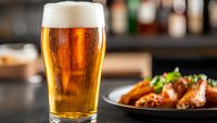 Cold glass of light beer with foam head and condensation drops next to spicy buffalo chicken wings on dark plate in bar setting, selective focus on beer.