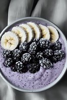 Purple smoothie bowl topped with fresh banana slices and frozen blackberries creates vibrant breakfast composition on gray background.