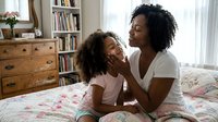 Adult woman of African descent with curly black hair wearing white t-shirt sitting on bed with young girl, both with curly hair, in bright bedroom with wooden furniture and bookshelves, daytime natural light.