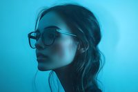 Young Asian woman wearing glasses in profile view against turquoise background, dramatic moody lighting creates atmospheric portrait with loose hair bun style.