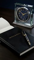 Crystal desk clock with navy blue face and gold accents beside black leather notebook and pen on dark wooden surface. Perfect for corporate office, business workspace, and professional environment styling.