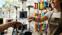 Female pet shop employee assisting customer with brown leather dog leash in store filled with colorful pet toys and supplies, demonstrating product quality and customer service in retail environment.