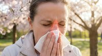 Middle-aged Caucasian woman sneezing into tissue outdoors during spring cherry blossom season, suffering from seasonal allergies and hay fever symptoms.