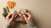 Hands tying golden ribbon bow on kraft paper gift box with dried orange slices and pine branch for festive holiday wrapping and celebration.