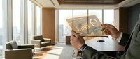 Professional businessman in suit holding stack of dollar bills in modern office with city skyline view through floor-to-ceiling windows and contemporary furniture.
