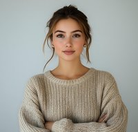 Young woman with natural makeup wearing beige knit sweater, arms crossed, looking at camera with gentle smile against light background. Professional portrait.