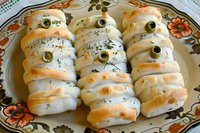 Spooky mummy-shaped baked pastries wrapped in strips of dough, decorated with green olives as eyes and herbs, served on floral plate for Halloween party snacks.