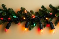 Festive string of multicolored Christmas lights with red, green, and orange bulbs illuminated against artificial pine garland on neutral background. Holiday decor.