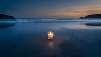 Glowing candle on wet sand beach at sunset with ocean waves and dramatic twilight sky creating peaceful meditation atmosphere for wellness and relaxation.