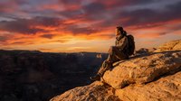 Hiker sitting on rocky cliff edge overlooking vast canyon landscape during dramatic golden sunset with orange and purple sky, contemplating nature and adventure.