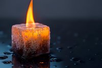 Glowing ice cube candle with orange flame on dark reflective surface with water drops, creating atmospheric mood lighting for spiritual practices.