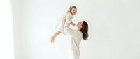 Joyful Caucasian mother lifting laughing toddler girl in white studio, celebrating love and family bond in bright minimal setting.