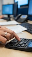 Businessman hand using calculator at office desk with computer monitors and documents for financial calculations and accounting work.
