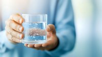 Glass of pure water with bubbles held in caucasian hands against light blue background, selective focus on transparent glass with fresh drinking water.