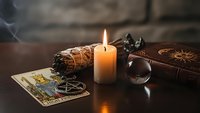 Mystical fortune telling setup with burning candle, tarot cards, crystal ball and sage bundle creating atmospheric divination scene for spiritual practices.