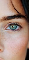 Close-up macro shot of young woman's bright blue-green eye with natural freckles on fair skin, showing detailed eyelashes and defined eyebrow against soft focus background.