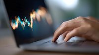 Close-up view of person typing on laptop keyboard with financial chart graph displayed on blurred screen, showing stock market trading and analysis.