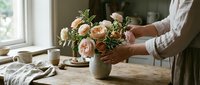 Woman arranging fresh peach roses in ceramic vase on rustic wooden table by window. Cozy home interior with natural light and pottery.