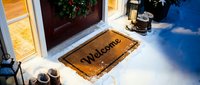 Welcome doormat at a snow-covered front door decorated with a Christmas wreath, warm glowing light, winter holiday home entrance scene.
