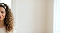 Young woman with long curly brown hair and olive skin tone smiling at camera in bright interior space, suitable for lifestyle and portrait use.