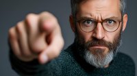 Mature bearded man in glasses pointing finger at camera with serious stern expression wearing green sweater against dark background. Professional portrait.