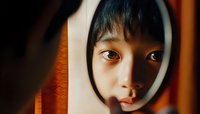 Young Asian woman looking through circular hole or frame, dramatic lighting creates mysterious mood with warm tones and shadows highlighting expressive eyes and face.