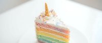 Slice of layered rainbow cake with golden unicorn horn topper, pastel frosting, and powdered sugar dusting on white plate for bakery, dessert, and celebration content.