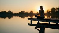 Silhouette of woman meditating in lotus pose on wooden dock at sunset over calm lake. Peaceful mindfulness and yoga practice for wellness and relaxation.