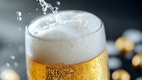 Cold beer glass with fresh foam head and splashing drops, condensation on surface against dark background, macro shot showing golden bubbles and texture.