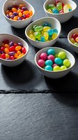 Colorful assorted candies in white bowls on dark slate, featuring gummy bears, jelly beans, and bubblegum balls in vibrant rainbow hues.
