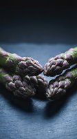 Fresh purple asparagus spears arranged on dark slate surface with moody lighting for culinary and healthy eating concepts.