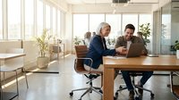 Two professional businessmen collaborating on laptop in bright modern office space with large windows and contemporary furniture for teamwork presentations.