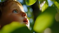 Young child gazing upward through green spring leaves with wonder and curiosity, face partially visible in soft natural sunlight creating magical atmosphere.