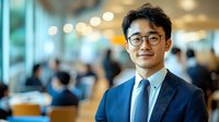 Young Asian businessman wearing glasses and navy suit smiling confidently in modern office environment with blurred colleagues in background. Professional corporate portrait.