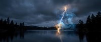 Dramatic lightning strike illuminates stormy sky over calm lake surrounded by dark forest silhouettes creating powerful natural spectacle.