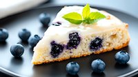 Slice of creamy blueberry cheesecake garnished with fresh mint leaf and whipped cream, served on black plate with scattered fresh blueberries, macro shot.
