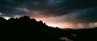Dramatic storm clouds gather over silhouetted mountains and river valley at sunset, creating moody atmospheric landscape for weather, nature, and environmental design projects.